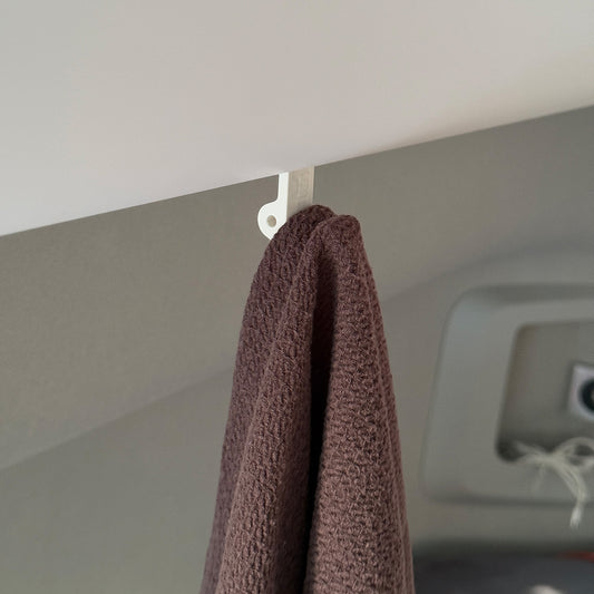 Ein robuster Multifunktionshaken in weiß, eingehängt an einem Schrank im VW Grand California. Am unteren Haken hängt eine Jacke, während durch die kleine Öse auf der Rückseite eine Schnur gezogen ist, an der ein Bluetooth-Lautsprecher befestigt wurde. Der Haken fügt sich unauffällig ins Fahrzeugdesign ein und bleibt sowohl bei geöffnetem als auch geschlossenem Schrank nutzbar. Perfekt für mehr Ordnung im Camper!