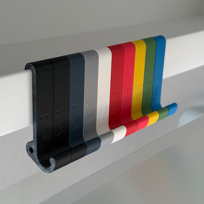 Ein robuster Multifunktionshaken in zehn unterschiedlichen Farben, eingehängt an einem Schrank im VW Grand California. Am unteren Haken hängt eine Jacke, während durch die kleine Öse auf der Rückseite eine Schnur gezogen ist, an der ein Bluetooth-Lautsprecher befestigt wurde. Der Haken fügt sich unauffällig ins Fahrzeugdesign ein und bleibt sowohl bei geöffnetem als auch geschlossenem Schrank nutzbar. Perfekt für mehr Ordnung im Camper!